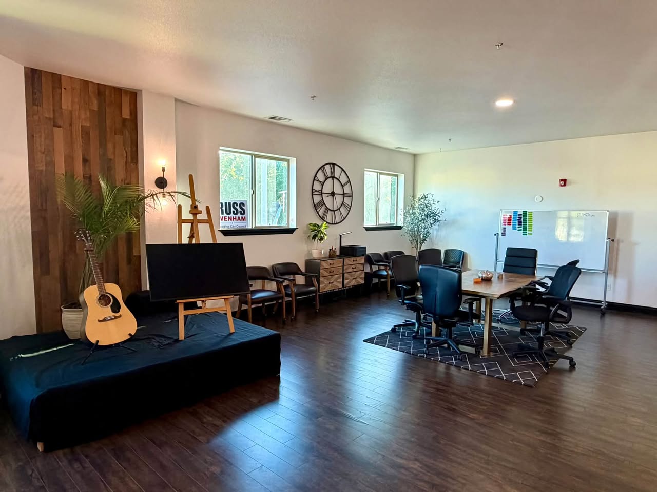 Conference table with ergonomic chairs, a rolling whiteboard, and a smart TV on an easel in a bright room with wood floors
